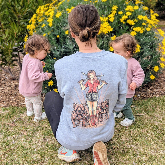 Woman in a beautiful light grey sweatshirt with a gorgeous design featuring twin pitties. She is in a garden bursting with floral blooms, with two children by her side.